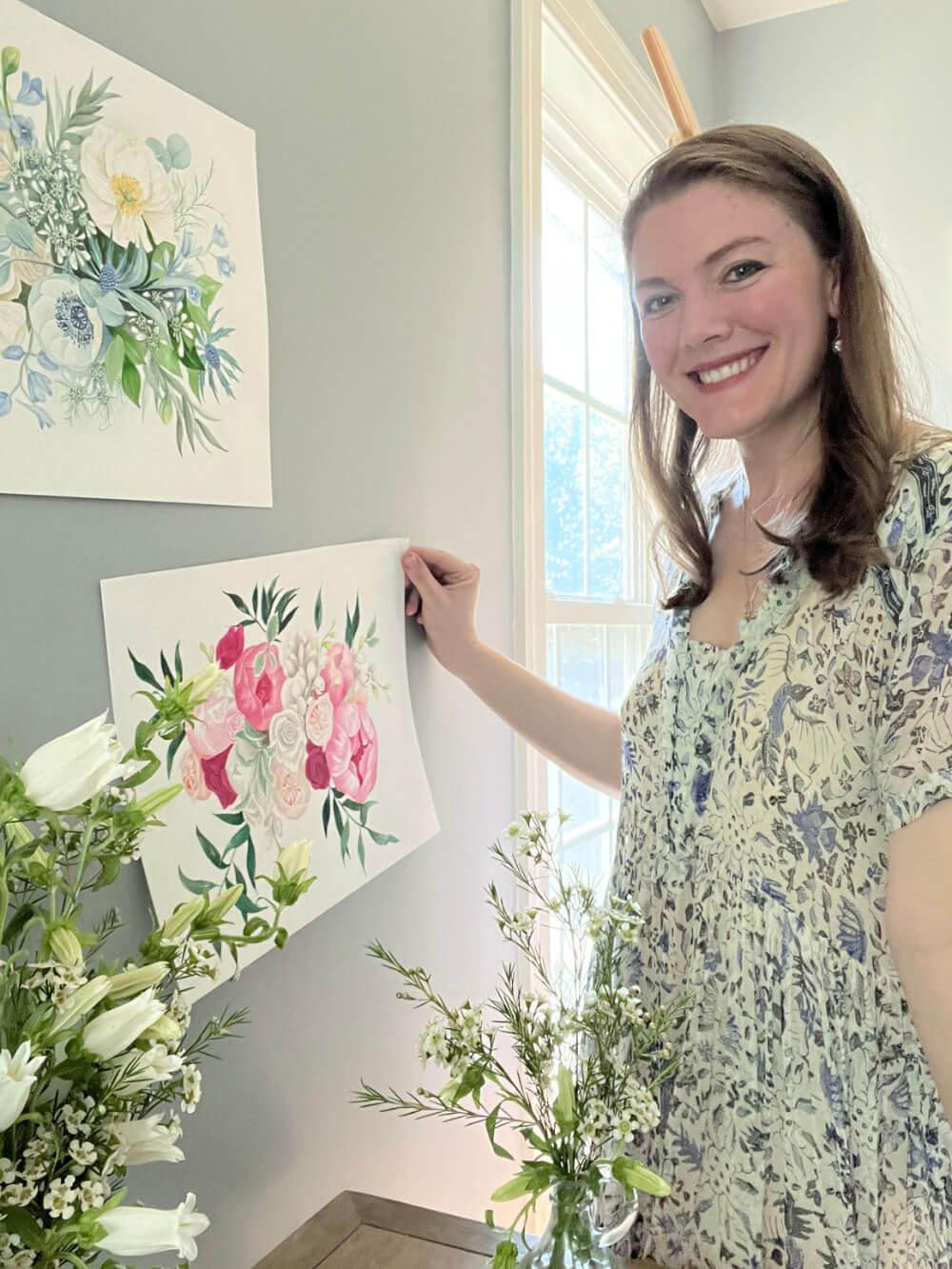 Courtney Hopkins smiling and standing in front of her pink and blue wedding bouquet paintings on the wall